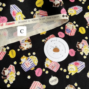 May include: Black fabric with a repeating pattern of cats holding popcorn boxes. The cats are wearing red and blue striped shirts and have yellow fur. The fabric has a white border with the text "of Animals designed by KOKKA Co.,Ltd made in Japan".