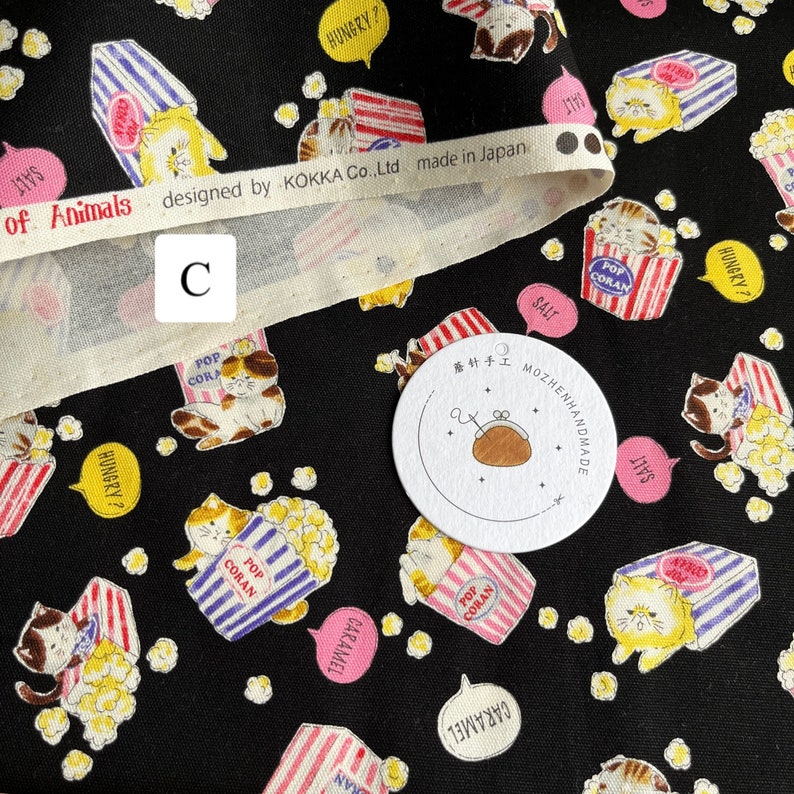 May include: Black fabric with a repeating pattern of cats holding popcorn boxes. The cats are wearing red and blue striped shirts and have yellow fur. The fabric has a white border with the text "of Animals designed by KOKKA Co.,Ltd made in Japan".