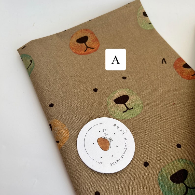 May include: Brown fabric with a repeating pattern of colorful bear faces. The fabric has black dots scattered throughout. A white circle with black text reads "HANDMADE MOZZIEHEN" in Japanese. The letter "A" is written in black on a white square.