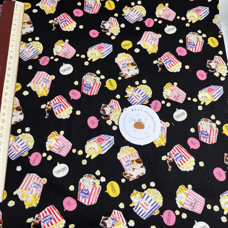 May include: Black fabric with a repeating pattern of cartoon cats holding boxes of popcorn. The boxes are red, white, and blue with the words "Pop Corn" on them. The cats are wearing different expressions and some have speech bubbles with words like "Hungry?" and "Caramel".