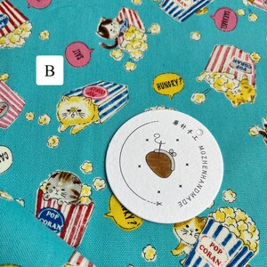 May include: A turquoise blue fabric with a repeating pattern of cats holding popcorn boxes. The boxes are red and white striped with the words "Pop Coran" printed on them. The cats are in various poses and have different facial expressions. There are also small white popcorn kernels scattered throughout the fabric. A white circle with black text reads "蘑针手工 MOZHENHANDMADE".