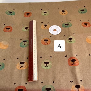 May include: A brown fabric with a repeating pattern of colorful bear faces. The fabric is textured and has a subtle sheen. The bear faces are in shades of green, orange, and brown. The fabric is measured with a ruler in inches.