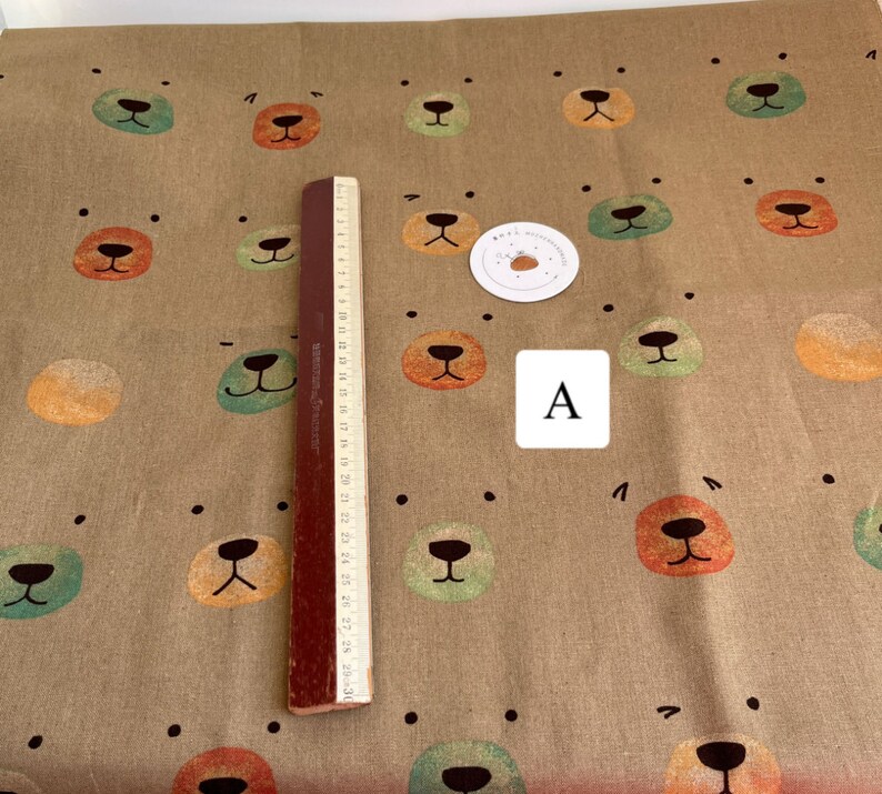 May include: A brown fabric with a repeating pattern of colorful bear faces. The fabric is textured and has a subtle sheen. The bear faces are in shades of green, orange, and brown. The fabric is measured with a ruler in inches.