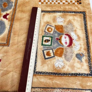 May include: A beige fabric with a brown, white, and blue patchwork design. The fabric features a whimsical illustration of a bird, a bee, and a snowman-like figure with a red hat. The fabric is measured with a ruler.