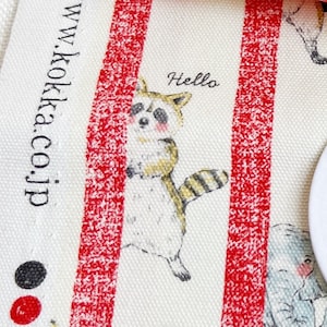 Könnte beinhalten: Weißer Stoff mit roten Streifen und einem Cartoon-Waschbären mit gelbem Schwanz. Der Waschbär hält sich an einem roten Streifen fest und hat einen rosa Rouge auf den Wangen. Der Text "Hello" ist über dem Waschbären geschrieben. Der Text "www.kokka.co.jp" ist auf der linken Seite des Bildes geschrieben.