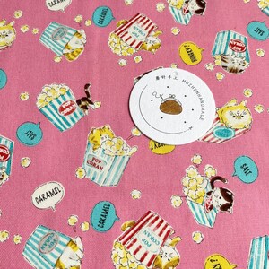 May include: Pink fabric with a repeating pattern of cats holding popcorn buckets. The buckets have red and white stripes and blue and white stripes. The cats are white with orange and black markings. The fabric has text that says "Pop Coran", "Salt", "Caramel", and "Hungry?" in a speech bubble. There is a white circle tag with black text that says "茶针手工 MOZHENHANDMADE".