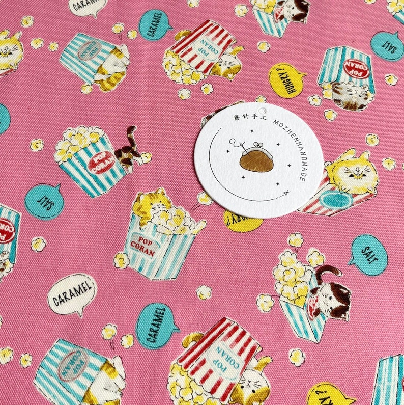 May include: Pink fabric with a repeating pattern of cats holding popcorn buckets. The buckets have red and white stripes and blue and white stripes. The cats are white with orange and black markings. The fabric has text that says "Pop Coran", "Salt", "Caramel", and "Hungry?" in a speech bubble. There is a white circle tag with black text that says "茶针手工 MOZHENHANDMADE".