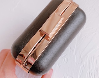 Può includere: Una pochette nera in legno con una chiusura in oro rosa. La pochette ha una finitura liscia e lucida.