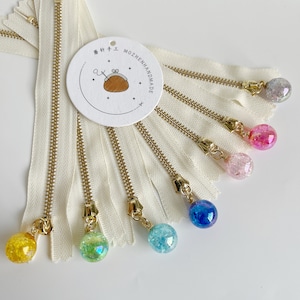 May include: A set of eight white zipper pulls with gold metal teeth and colorful glass beads. The beads are iridescent and have a speckled appearance. The zipper pulls are labeled "MOZHENHANDMADE".