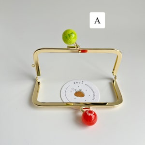 May include: Gold-toned metal frame with a green and red glass bead charm. The frame is for a small purse or wallet. The frame is on a white surface. The text on the tag reads "MOZHENHANDMADE".