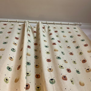May include: A pair of cream-colored curtains with a repeating pattern of brown, green, and orange bear faces. The curtains are hung on a white curtain rod.