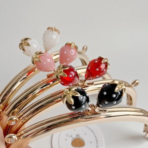 Op de afbeelding: Vier goudkleurige metalen handtaschframes met verschillende gekleurde aardbei-vormige sluitingen. De sluitingen zijn rood, roze, wit en zwart met witte stippen.