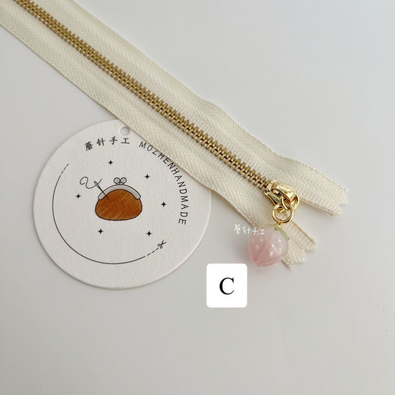 May include: A white zipper with a gold slider and a pink heart-shaped charm. The charm has a gold loop and a small gold bead. The zipper is labeled "MOZHENHANDMADE" and "C".
