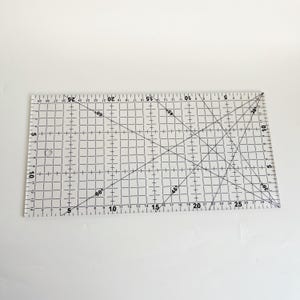 Può includere: Un righello in plastica trasparente con un motivo a griglia e linee inclinate. Il righello è graduato in pollici e include segni di gradi per 30, 45, 60 e 90 gradi.