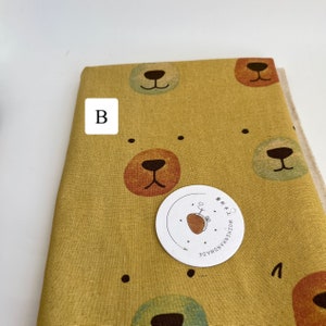May include: Yellow fabric with a repeating pattern of brown, orange, and green bear faces. The fabric has a white label with the text "MOZHEH.NOMADE Handmade" in black.
