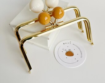 May include: Two gold metal purse frames with white and yellow round beads attached. The frames are on a white surface with a white tag that says "MOZHENHANDMADE" and has a small illustration of a purse.