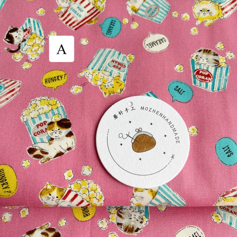 May include: Pink fabric with a repeating pattern of cats holding popcorn boxes. The boxes have blue and white stripes and say "Pop Coran" on them. There are also speech bubbles that say "Hungry?", "Salt", and "Caramel". A white circle tag with black text says "蘑针手工 MOZHENHANDMADE".