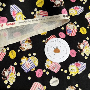 May include: Black fabric with a repeating pattern of cartoon cats holding boxes of popcorn. The boxes are red and white striped with the words "Pop Coran" on them. The cats are wearing blue and white striped shirts. The fabric has a white border with the text "of Animals designed by KOKKA Co.,Ltd made in Japan".