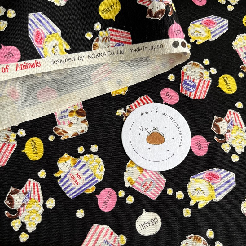 May include: Black fabric with a repeating pattern of cartoon cats holding boxes of popcorn. The boxes are red and white striped with the words "Pop Coran" on them. The cats are wearing blue and white striped shirts. The fabric has a white border with the text "of Animals designed by KOKKA Co.,Ltd made in Japan".