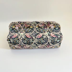 Può includere: Una pochette rettangolare con un motivo floreale e di uccelli nei toni del nero, rosa, verde e giallo. La pochette ha un bordo di pompon e due chiusure con bottoni rosa chiaro. Il design è dettagliato.