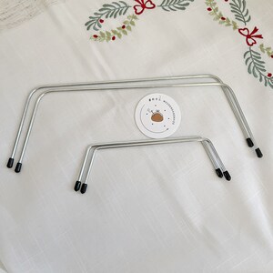 May include: Two silver metal cake cooling racks with black plastic feet. The racks are on a white tablecloth with a small white circle sticker with the text "MOTHERMADE" and a small brown illustration of a cake.