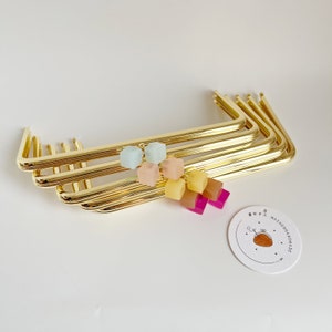 Può includere: Cinque telai per borse in metallo dorato impilati uno sopra l'altro. I telai sono decorati con quadrati di resina colorati in tonalità di rosa, giallo e blu. Un piccolo cartellino bianco con il testo "MOZHENIA HANDMADE" si trova nell'angolo in basso a destra dell'immagine.
