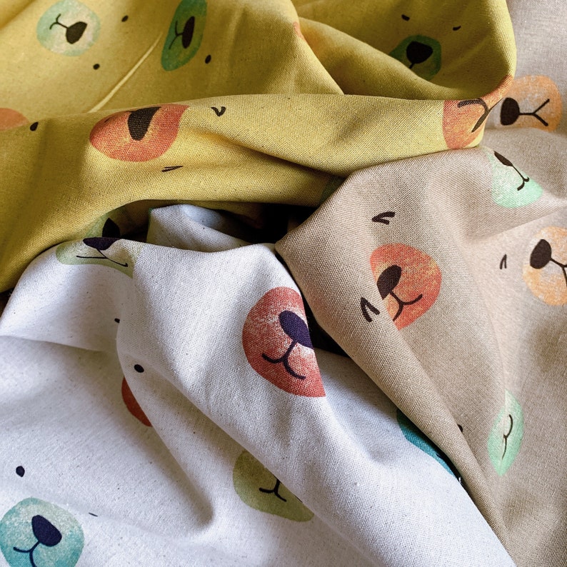 May include: Three fabric swatches with different colored backgrounds and bear face patterns. The first swatch is yellow with a brown bear face, the second is beige with a red bear face, and the third is white with a green bear face.