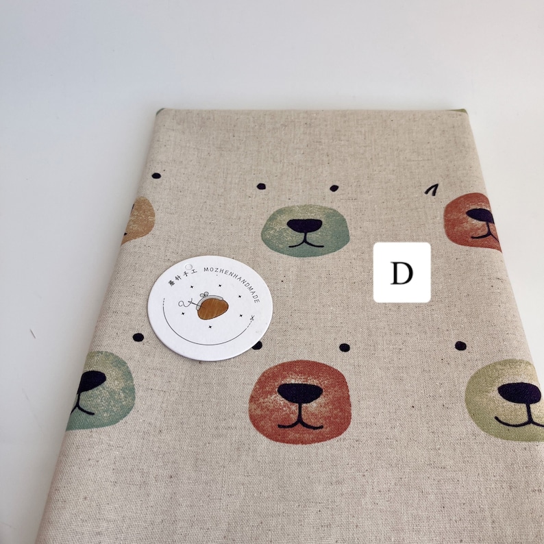 May include: Beige fabric with a repeating pattern of colorful bear faces. The fabric has black dots scattered throughout. A white circle label with the text "MOZHENHANDMADE" is attached to the fabric. The letter "D" is written in black on a white square.