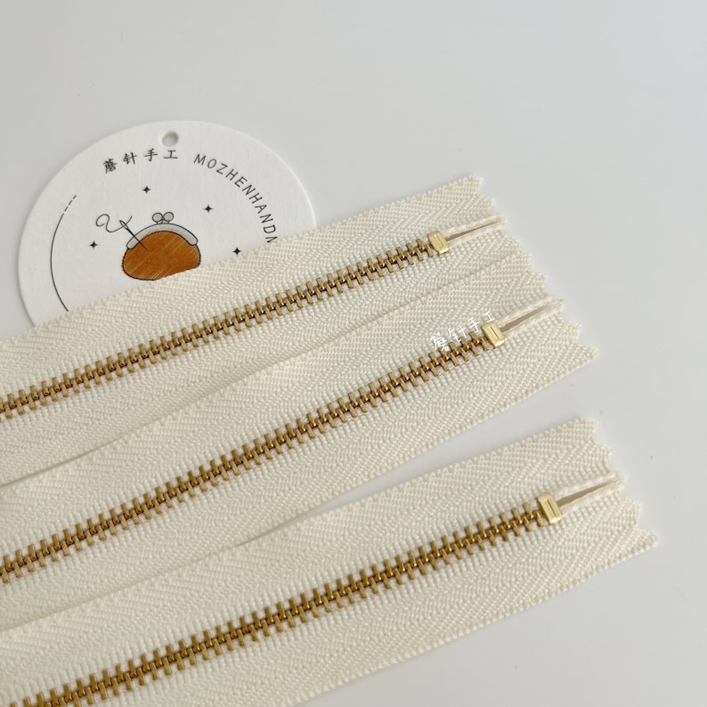 May include: Three white zipper tapes with gold metal teeth. The zipper tapes are laid out in a row, with the top tape slightly overlapping the middle tape, and the middle tape slightly overlapping the bottom tape.