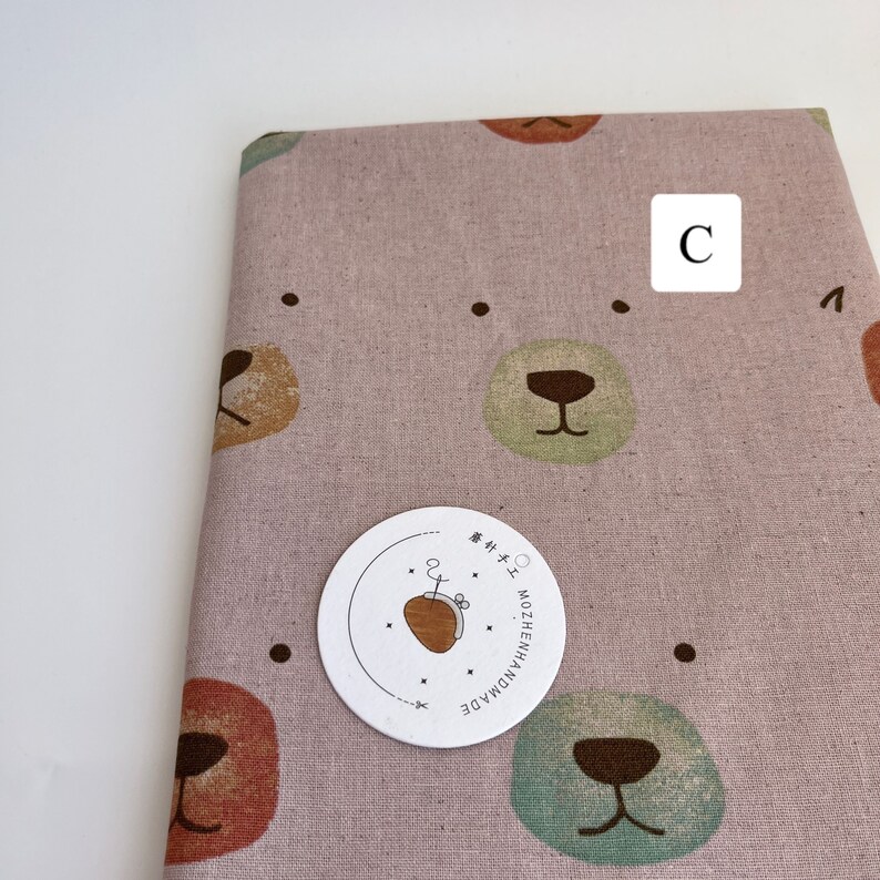 May include: Pink fabric with a repeating pattern of brown bear faces. The fabric has a subtle texture and is perfect for crafting projects. The fabric is labeled "HANDMADE NOZZHENH" in Japanese.