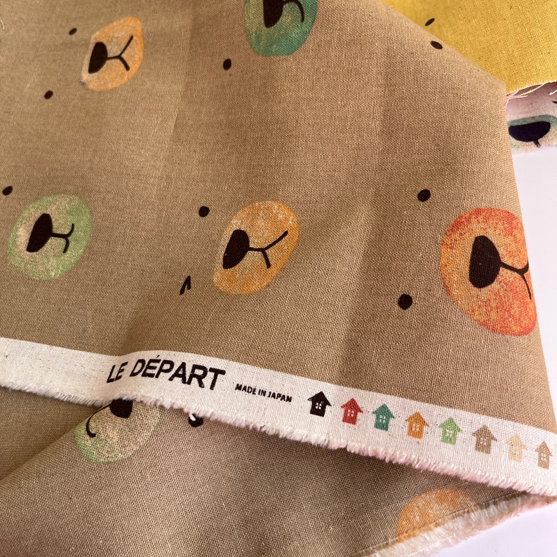 May include: A brown fabric with a repeating pattern of colorful cartoon faces. The fabric has a white label with the text "LE D&Eacute;PART MADE IN JAPAN".
