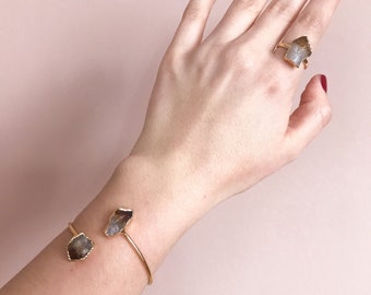 May include: A gold bracelet with two crystal accents. The bracelet is on a person's wrist. The person is also wearing a gold ring with a crystal accent.