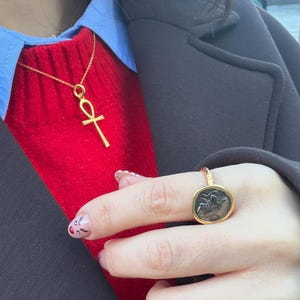 May include: A gold-toned necklace featuring an ankh pendant on a delicate chain, paired with a gold-toned ring that has a circular, dark-colored coin-style centerpiece with an embossed design, worn over a red sweater, blue shirt, and dark coat.