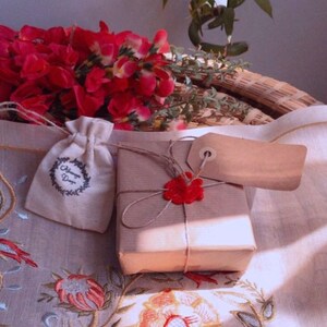 K&ouml;nnte beinhalten: Eine kleine, braune Geschenkbox, die in braunes Papier eingewickelt ist und mit einem roten Wachssiegel und einer Schnur verziert ist. Die Box steht auf einer wei&szlig;en Tischdecke mit einem Blumenmuster. Neben der Box liegt ein kleiner, wei&szlig;er Stoffbeutel mit dem Text "Always Dear".