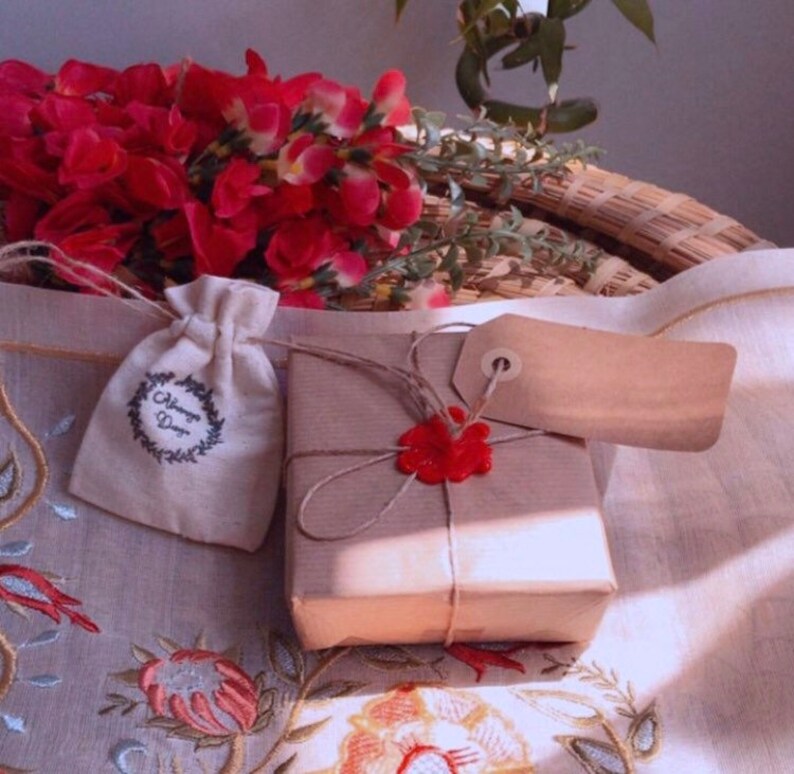 K&ouml;nnte beinhalten: Eine kleine, braune Geschenkbox, die in braunes Papier eingewickelt ist und mit einem roten Wachssiegel und einer Schnur verziert ist. Die Box steht auf einer wei&szlig;en Tischdecke mit einem Blumenmuster. Neben der Box liegt ein kleiner, wei&szlig;er Stoffbeutel mit dem Text "Always Dear".