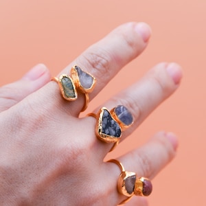 May include: Three gold rings with different colored stones. The rings are on a finger. The stones are green, black and white, and red.
