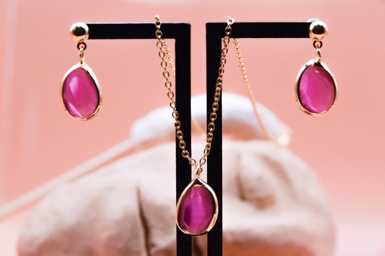 May include: A gold chain necklace and earrings with a teardrop-shaped pink stone pendant and matching earrings.