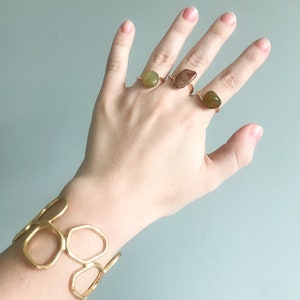 May include: A hand wearing three gold rings with different shaped green and brown stones. The hand is also wearing a gold bracelet with a geometric design.