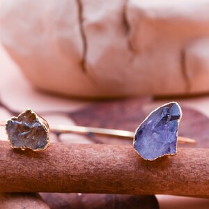 Puede incluir: Una pulsera de oro con dos piedras de cristal en bruto. Las piedras son de color marr&oacute;n claro y morado. La pulsera est&aacute; apoyada sobre una superficie de madera marr&oacute;n.