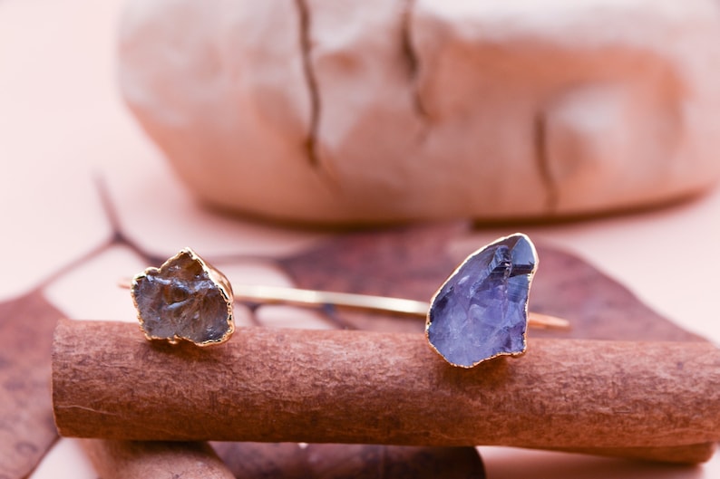 Puede incluir: Una pulsera de oro con dos piedras de cristal en bruto. Las piedras son de color marr&oacute;n claro y morado. La pulsera est&aacute; apoyada sobre una superficie de madera marr&oacute;n.