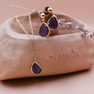 May include: A gold necklace and earrings with a teardrop-shaped purple gemstone pendant. The jewelry is displayed in a small clay dish with the word "LMAMIXA" inscribed on the side.