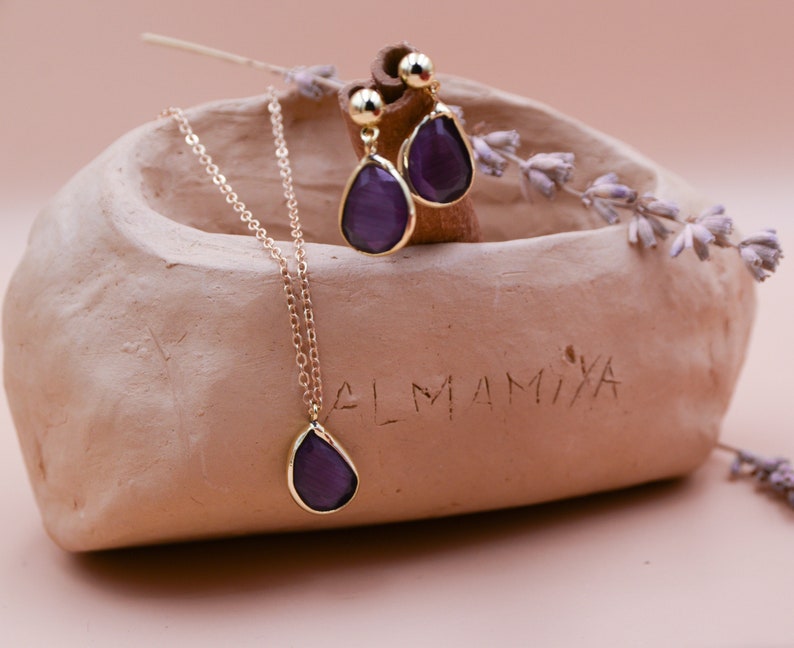 May include: A gold necklace and earrings with a teardrop-shaped purple gemstone pendant. The jewelry is displayed in a small clay dish with the word "LMAMIXA" inscribed on the side.