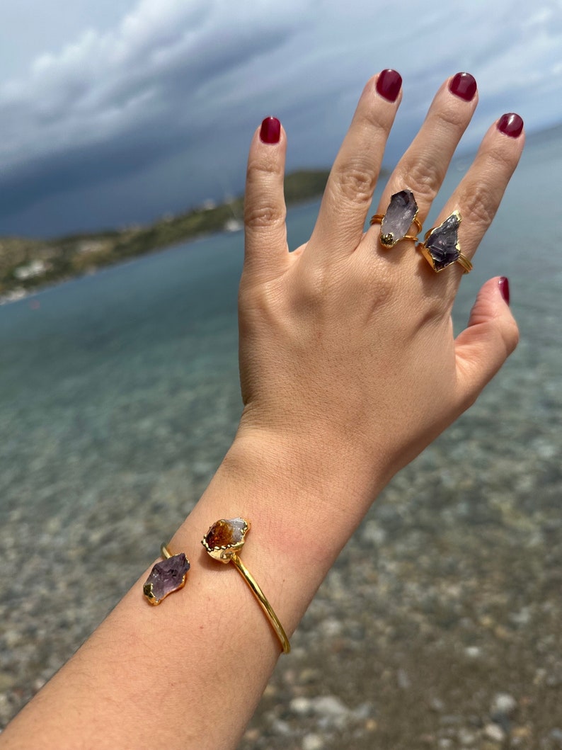 Puede incluir: Anillos de oro con piedras de amatista y citrino. Los anillos se llevan en una mano con esmalte de u&ntilde;as rojo. La mano tambi&eacute;n lleva una pulsera de oro con piedras de amatista y citrino.