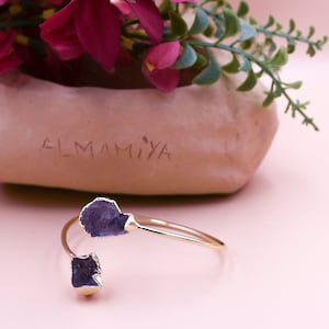 Puede incluir: Un brazalete de oro con dos piedras de amatista p&uacute;rpura. El brazalete est&aacute; sobre una superficie rosa.