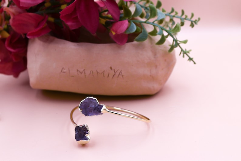 Puede incluir: Un brazalete de oro con dos piedras de amatista p&uacute;rpura. El brazalete est&aacute; sobre una superficie rosa.