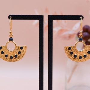 K&ouml;nnte beinhalten: Ein Paar goldene Ohrringe mit einem Halbkreisdesign und schwarzen Akzenten. Die Ohrringe h&auml;ngen an einem schwarzen St&auml;nder.