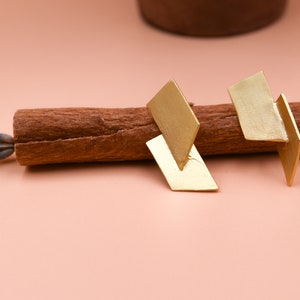 May include: A pair of gold geometric earrings with a modern design. The earrings are made of two overlapping rectangles, creating a unique and stylish look.