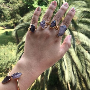 Puede incluir: Una mano con cinco anillos de oro con piedras de amatista. Cada anillo presenta una piedra de amatista de forma diferente.