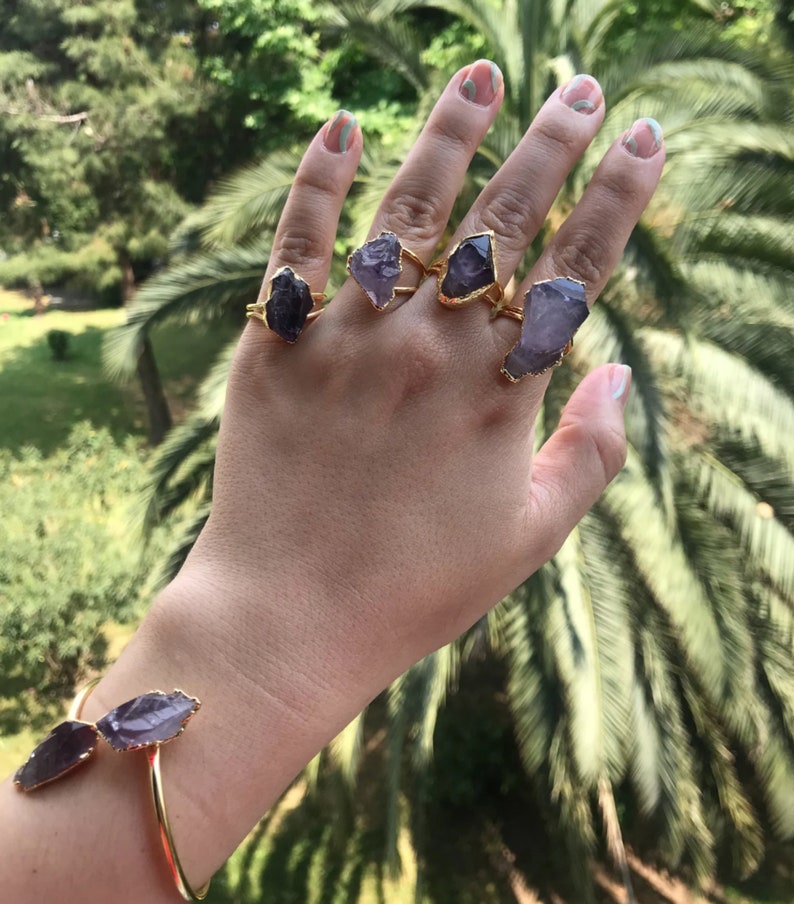 Puede incluir: Una mano con cinco anillos de oro con piedras de amatista. Cada anillo presenta una piedra de amatista de forma diferente.