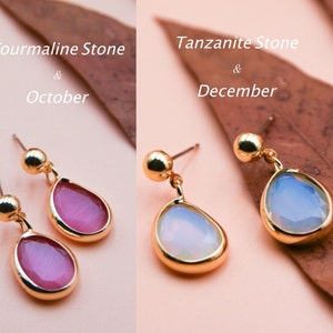 May include: Two pairs of gold earrings with teardrop-shaped stones. The earrings on the left have pink stones and the text "Tourmaline Stone & October". The earrings on the right have light blue stones and the text "Tanzanite Stone & December".